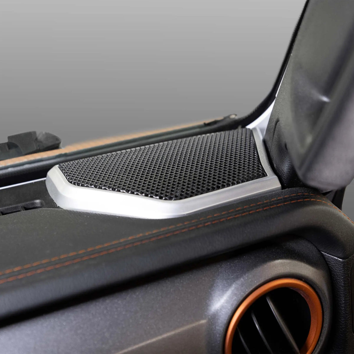 Installed dashboard-corner speaker grille with silver trim beside an orange-ringed air vent