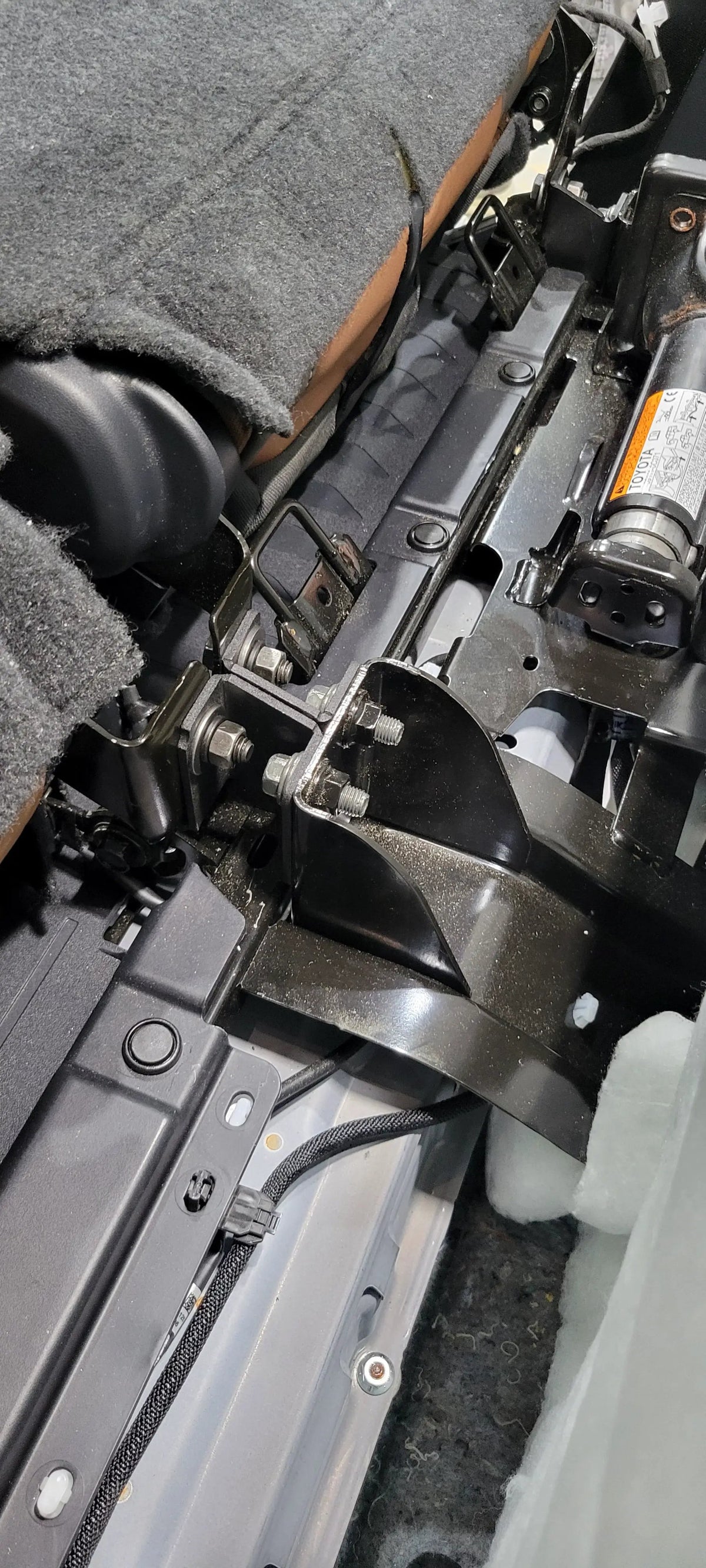 Close-up of brackets and bolted supports behind a raised rear seat beside the factory jack