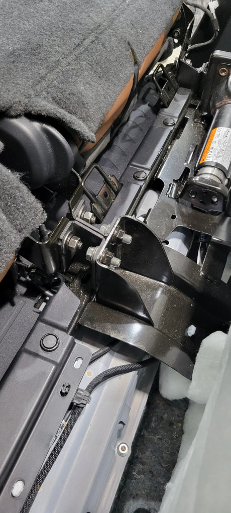 Close-up of brackets and bolted supports behind a raised rear seat beside the factory jack
