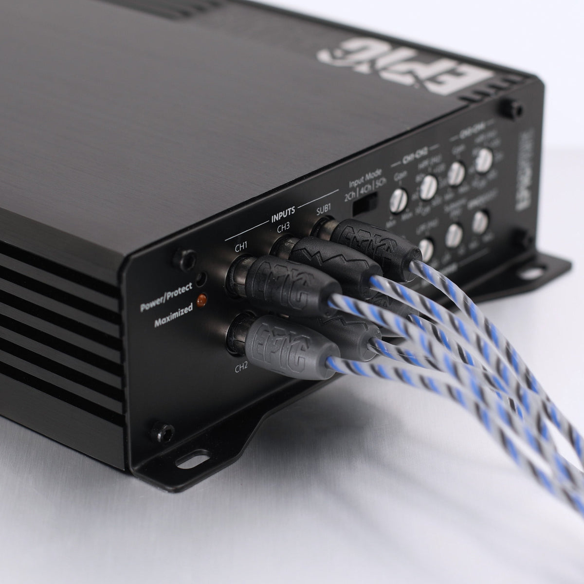 A close-up of a black audio power amplifier or receiver with blue-and-white braided cables plugged into its front panel, photographed on a light neutral background.