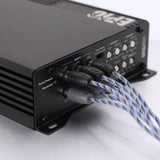 A close-up of a black audio power amplifier or receiver with blue-and-white braided cables plugged into its front panel, photographed on a light neutral background.