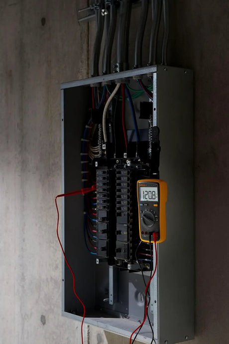 Yellow digital multimeter hanging beside an open breaker panel, with red and black test leads connected