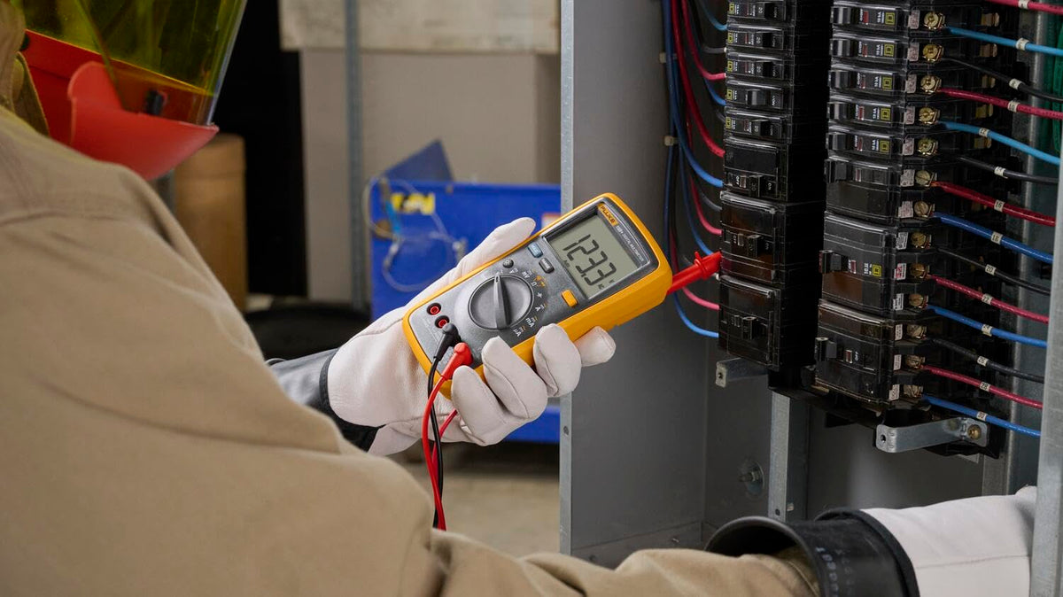 Side view of a technician holding a digital multimeter and testing an open circuit-breaker panel