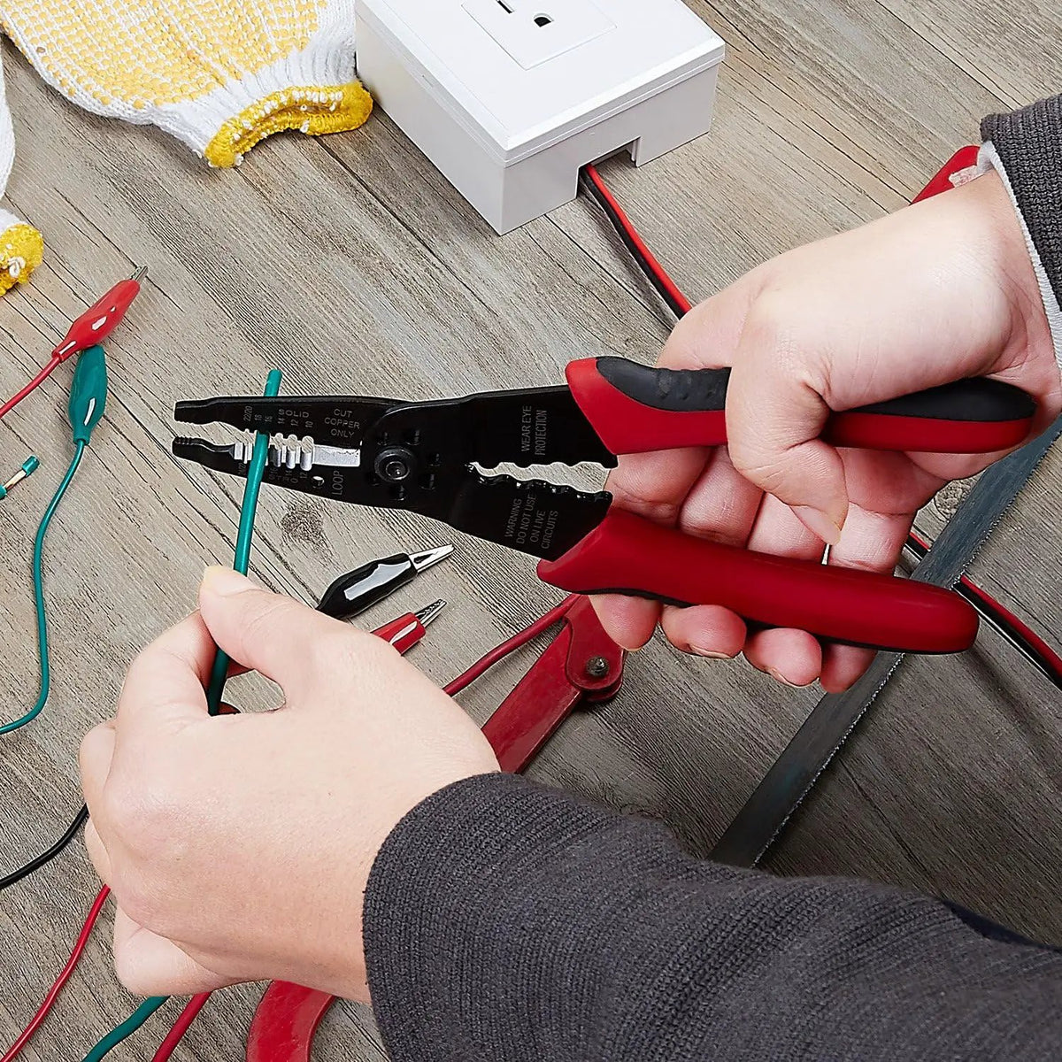 Hands using the red-and-black wire stripper on a green wire at a workbench