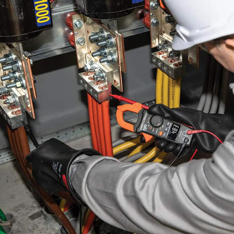 Technician using an orange-jawed Klein clamp meter and test probe at an electrical busbar assembly
