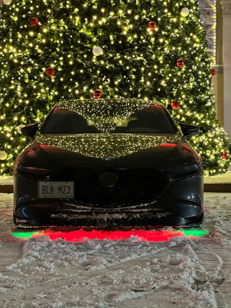 Black car with red and green underglow parked in snow beside a lighted tree