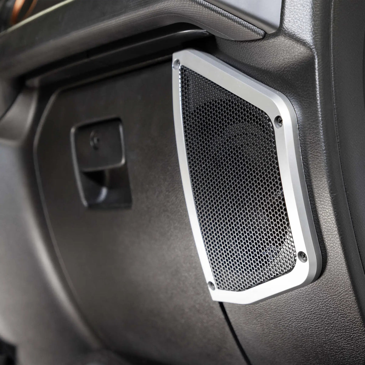 Rectangular speaker grille with silver trim installed in black vehicle interior paneling